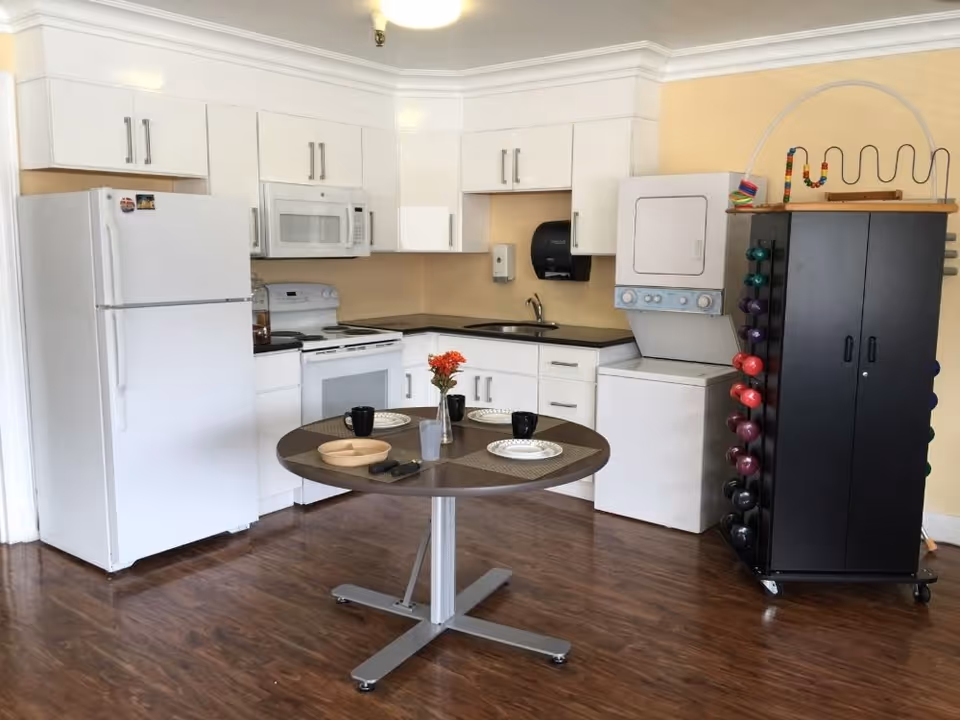A clean kitchen area with white cabinets, a white refrigerator, stove, microwave, and a stacked washer and dryer unit. In the center, there is a round table set with plates, cups, and a small vase with red flowers. To the right, there is a black cabinet with colorful dumbbells and a bead maze toy on top. The floor is wooden and the walls are painted light yellow.