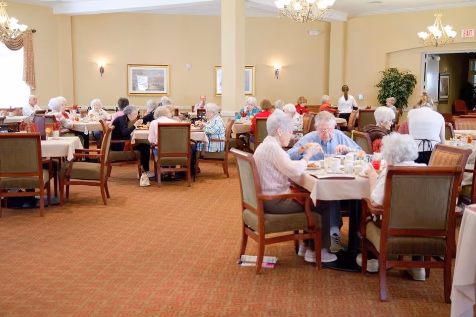 A dining room in a senior living facility with elderly residents seated at tables eating and socializing. The room has beige walls, framed artwork, chandeliers, and large windows with curtains. Staff members are seen assisting residents.