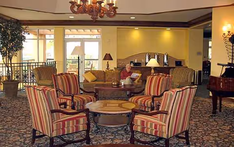 A comfortable hotel-like senior living lounge with striped chairs around a central table; a resident reading.
