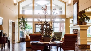 A bright and spacious living room with high ceilings and large windows allowing natural light to fill the space. The room features comfortable upholstered chairs around a central wooden coffee table with a large plant centerpiece. There is a fireplace on the right side with a framed artwork above it, and additional seating and plants are visible in the background.