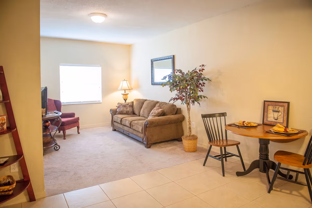 A cozy living room area with a brown sofa, a floor lamp, a framed mirror on the wall, a potted plant, and a small round wooden dining table set with two chairs and place settings. The room has beige walls, a window with blinds, and a carpeted floor transitioning to tiled flooring near the dining area.