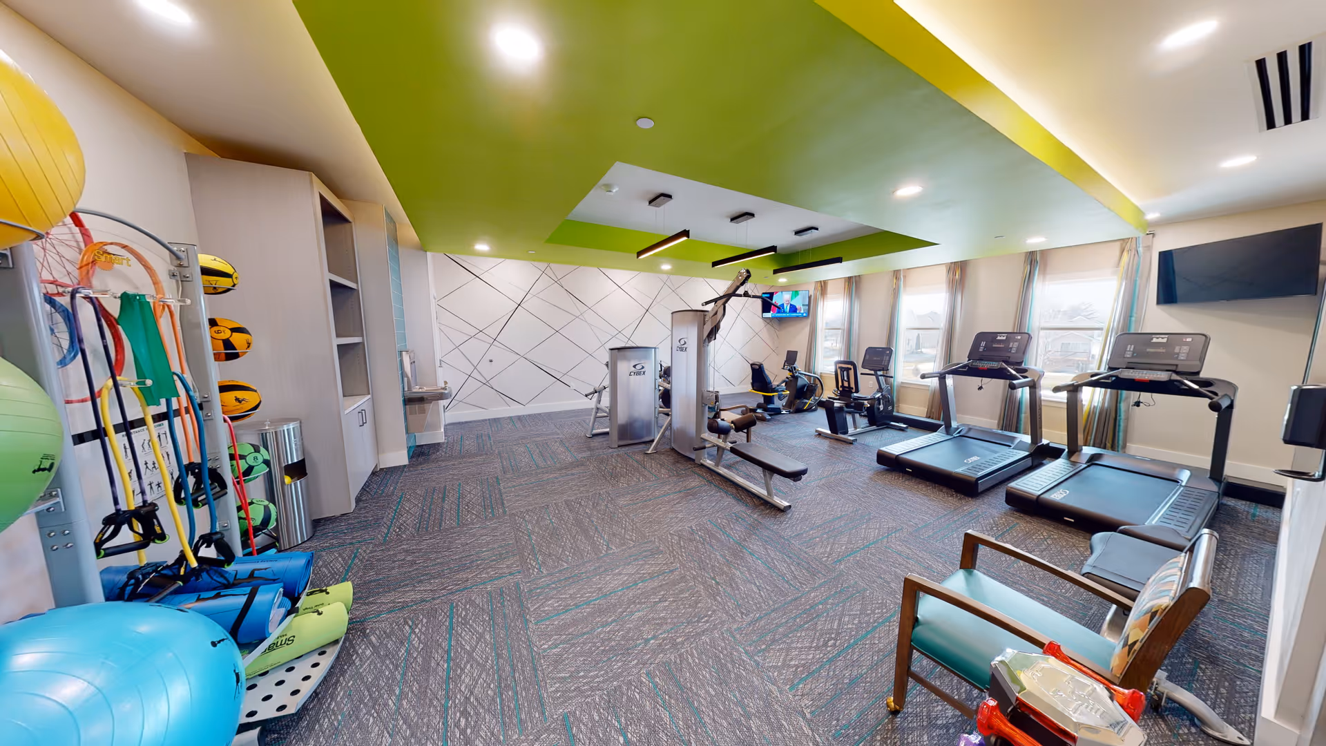 A bright and spacious fitness room in CedarStone Senior Living featuring exercise equipment including treadmills, stationary bikes, a multi-functional weight machine, and various fitness accessories like exercise balls, resistance bands, and medicine balls. The room has large windows allowing natural light, a green accent ceiling, and a wall-mounted TV.