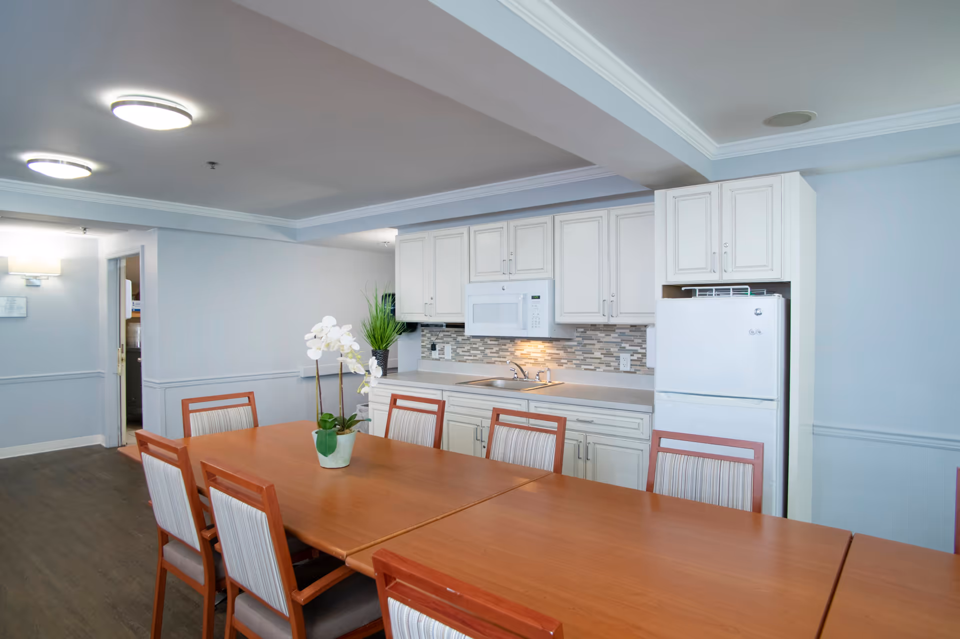 A bright and clean kitchen and dining area in a senior living facility. The room features a long wooden dining table with eight chairs, a white refrigerator, white cabinets, a microwave, a sink, and a decorative plant on the table. The walls are painted light blue with white trim, and the floor is dark wood.