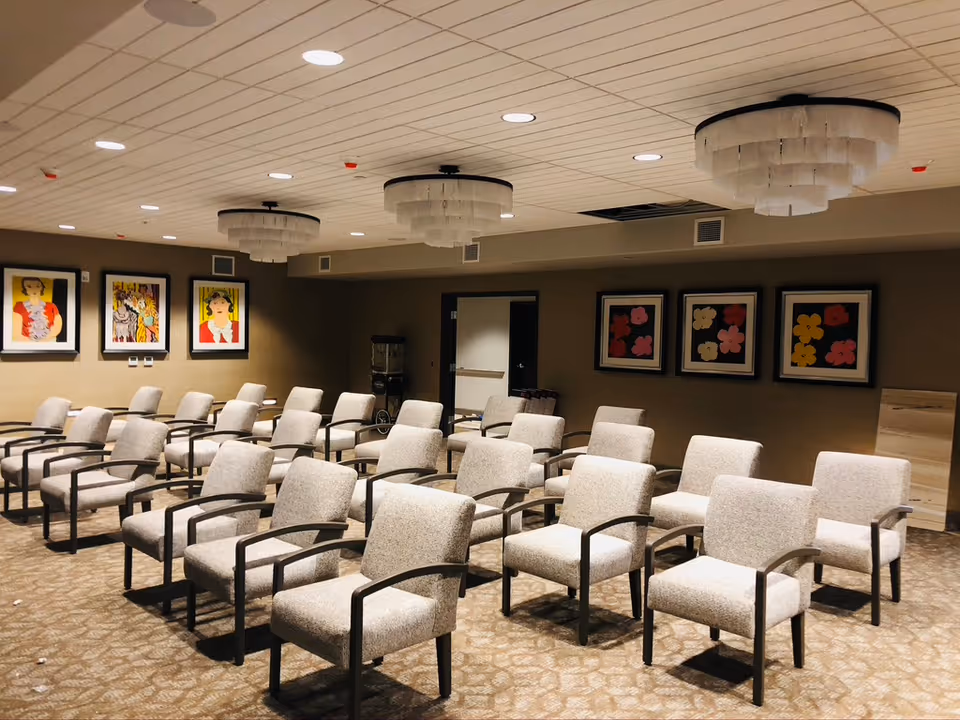 A room with multiple rows of beige upholstered chairs with dark armrests arranged facing forward. The ceiling has recessed lighting and three large, round chandeliers with layered white shades. The walls are decorated with framed colorful artwork, including portraits and floral designs. The carpet has a patterned design, and there is a water dispenser in the back corner near a door.