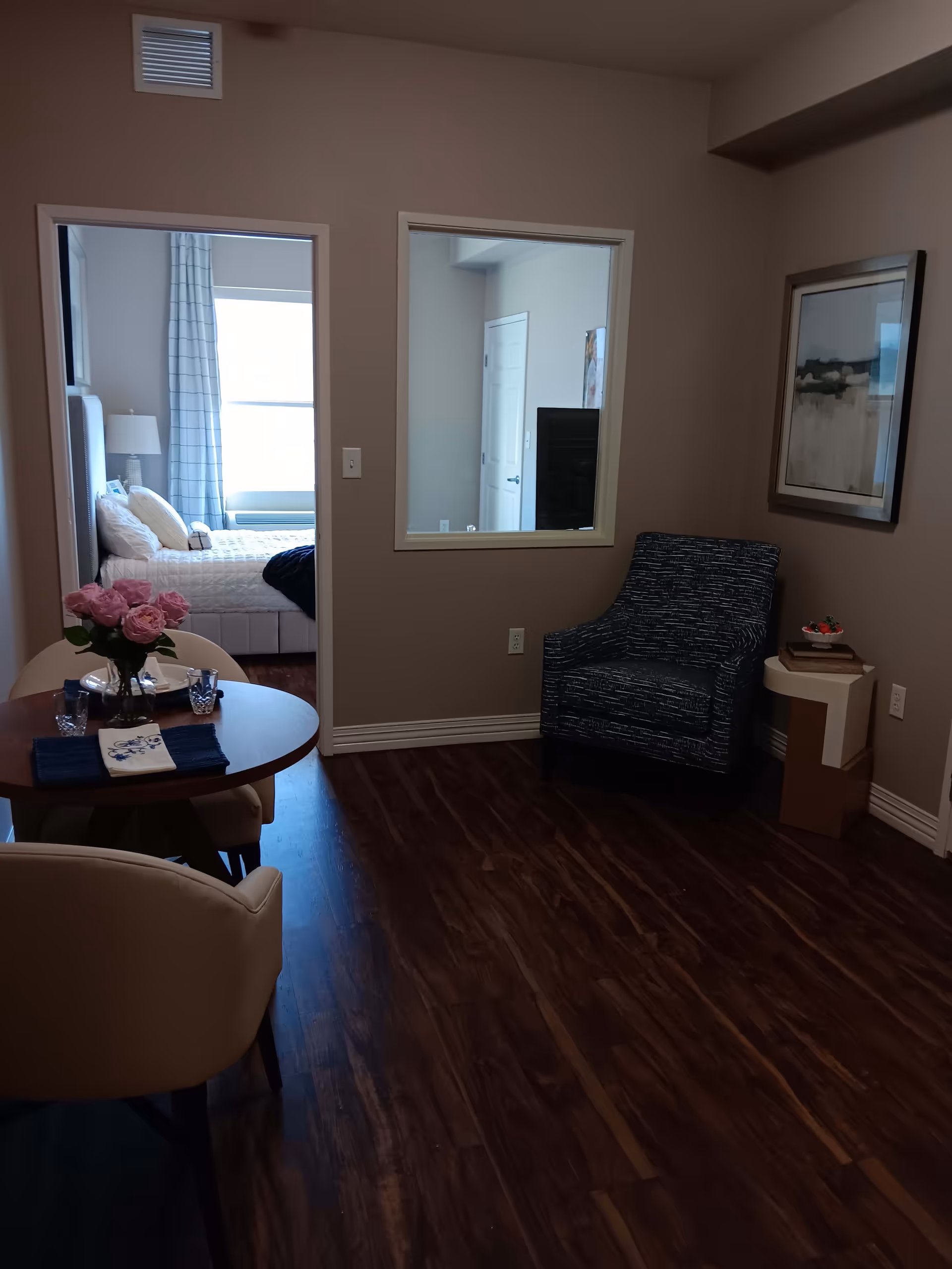 Interior view of a senior living facility room with dark wood flooring. There is a small round dining table with two beige chairs and a vase of pink flowers on the table. To the right, there is a blue patterned armchair next to a small side table with books and a decorative item. A framed abstract painting hangs on the wall above the armchair. Through an open doorway, a bedroom is visible with a bed, white bedding, pillows, a bedside lamp, and a window with curtains.