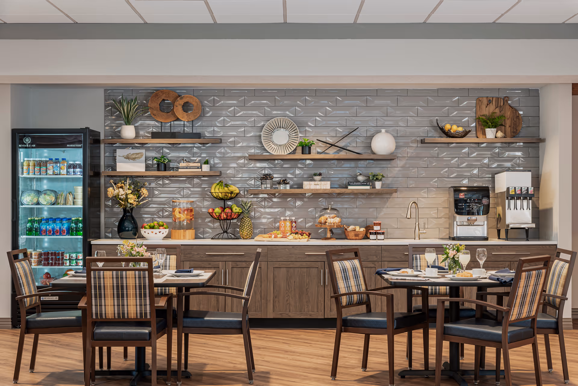 A dining area with two round tables set with plates, glasses, and napkins. Behind the tables is a countertop with wooden cabinets, decorated with fruit baskets, jars, a water dispenser, and shelves holding decorative items and plants. A refrigerator stocked with beverages is on the left side.