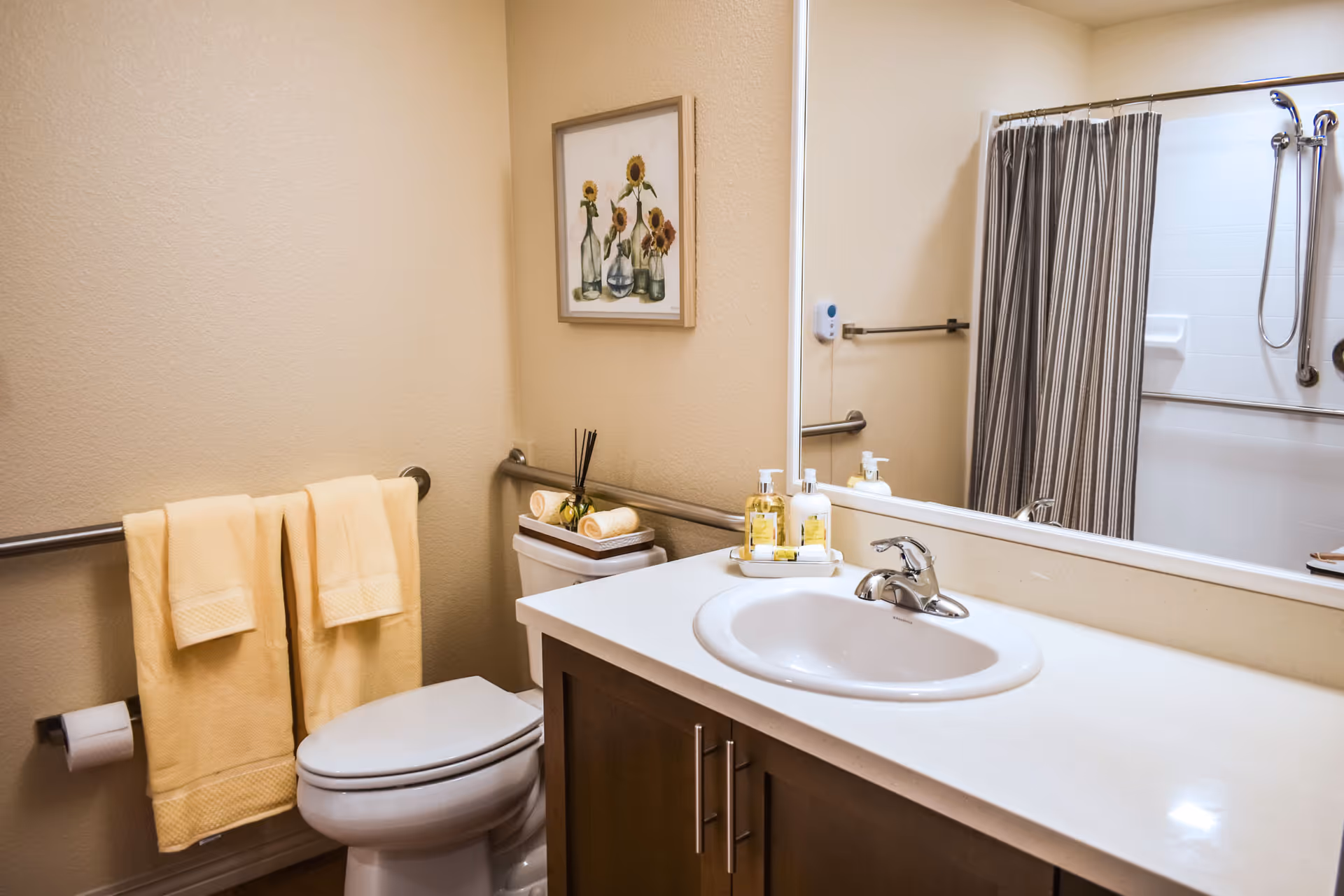 A clean and well-lit bathroom featuring a white toilet with two yellow towels hanging on a metal bar above the toilet paper holder. A white countertop with a sink and silver faucet is visible, along with soap dispensers and a decorative tray. A large mirror reflects a shower area with a striped shower curtain and grab bars. A framed picture of sunflowers in vases hangs on the beige wall above the toilet.