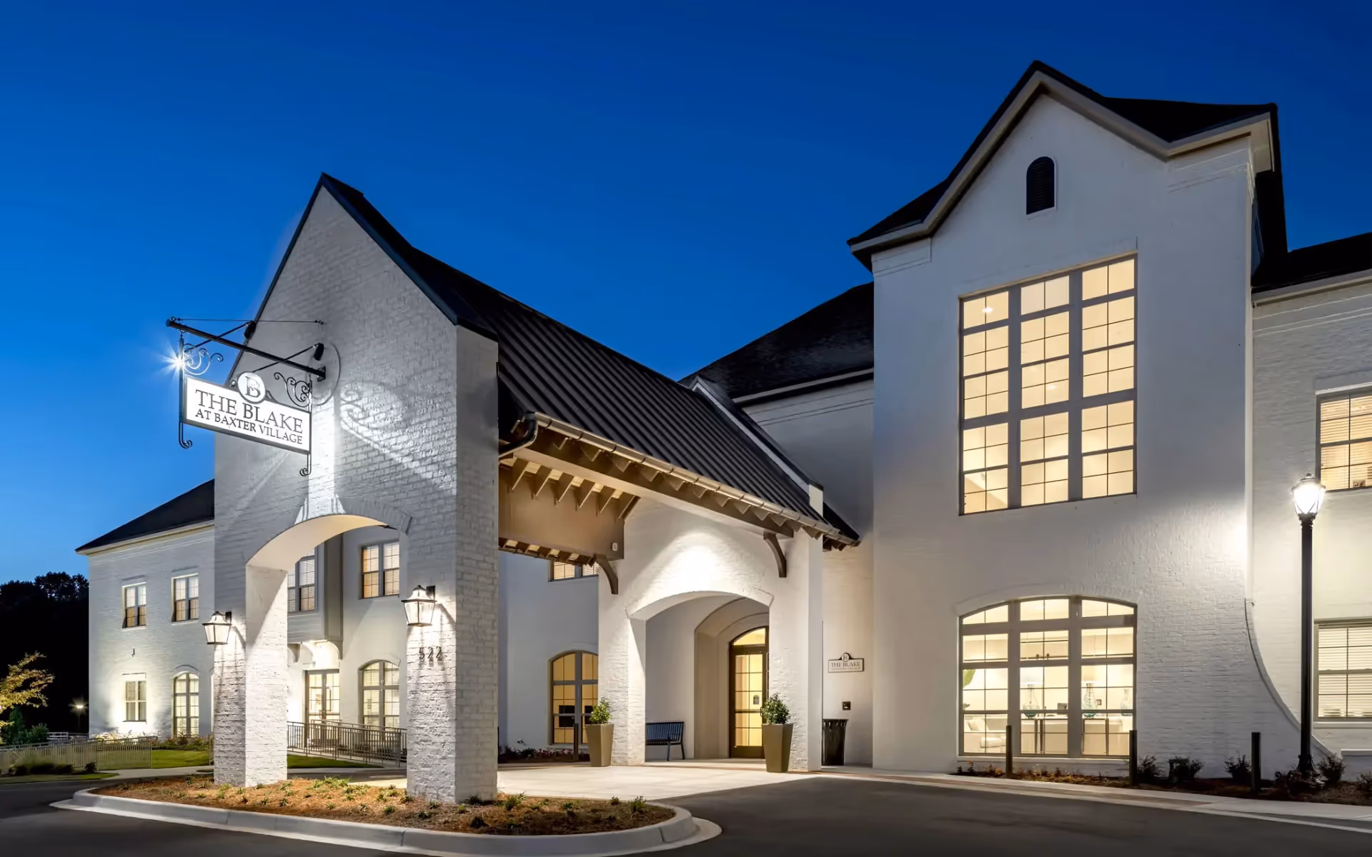 Exterior view of The Blake at Baxter Village senior living facility at dusk, showing a white brick building with large windows illuminated from inside, a covered entrance with a black metal roof, and a sign displaying the facility's name.