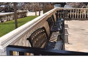 Outdoor patio area with a row of black metal benches along a stone balustrade overlooking a grassy area and trees.