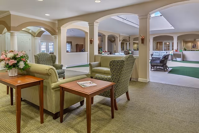 A spacious and well-lit common area in a senior living facility featuring green upholstered armchairs and sofas arranged around wooden side tables. The room has beige walls with white trim, arched openings, and a skylight allowing natural light to enter. There is a floral arrangement on one of the tables and additional seating visible in the background.