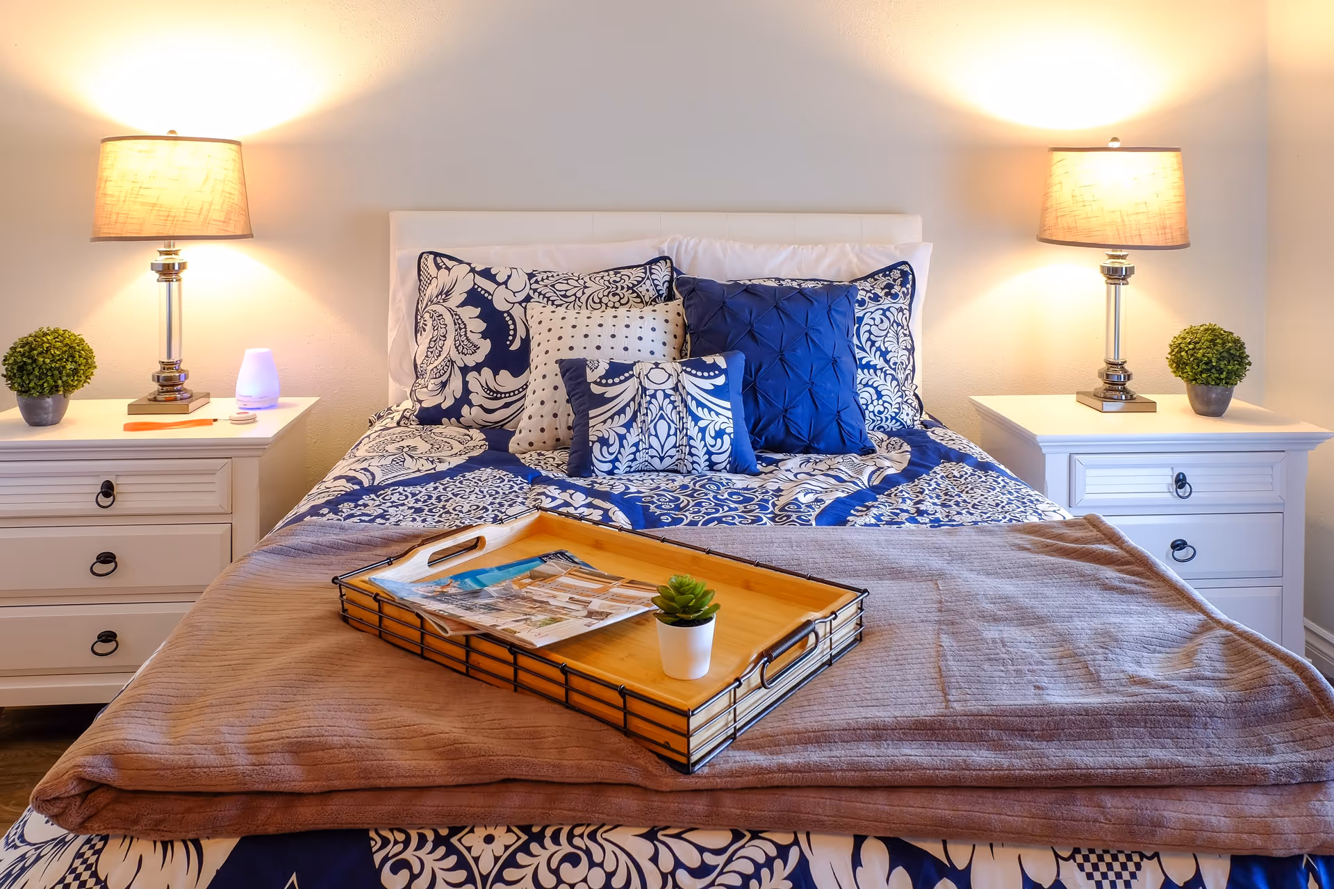Well-lit bedroom with a neatly made bed featuring blue patterned bedding, bedside tables with lamps, and a wooden tray on the bed.