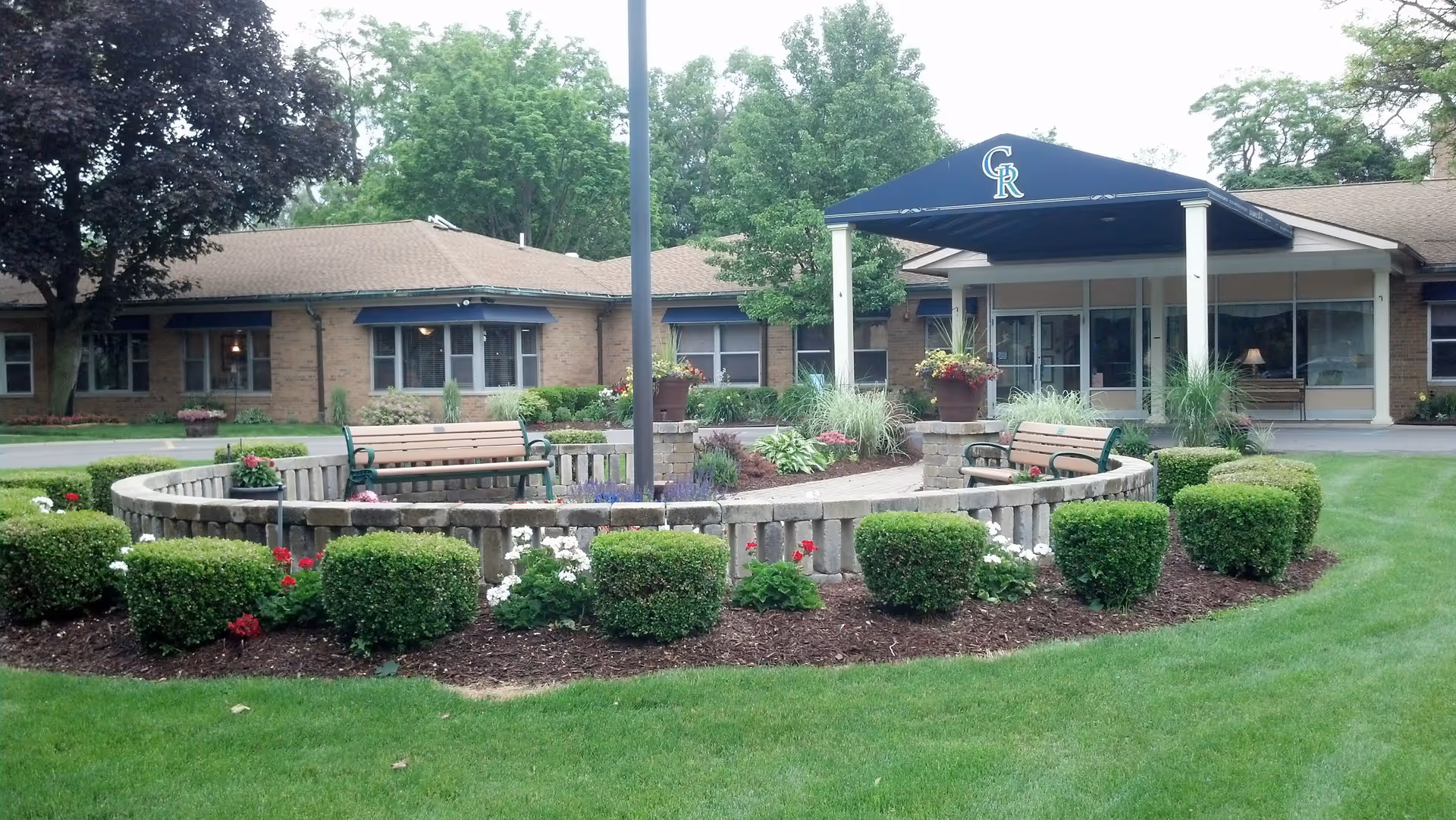 Outdoor garden area in front of a single-story brick building with a covered entrance. The garden features neatly trimmed bushes, flower beds, a circular stone wall, and wooden benches. Trees and well-maintained grass surround the area.