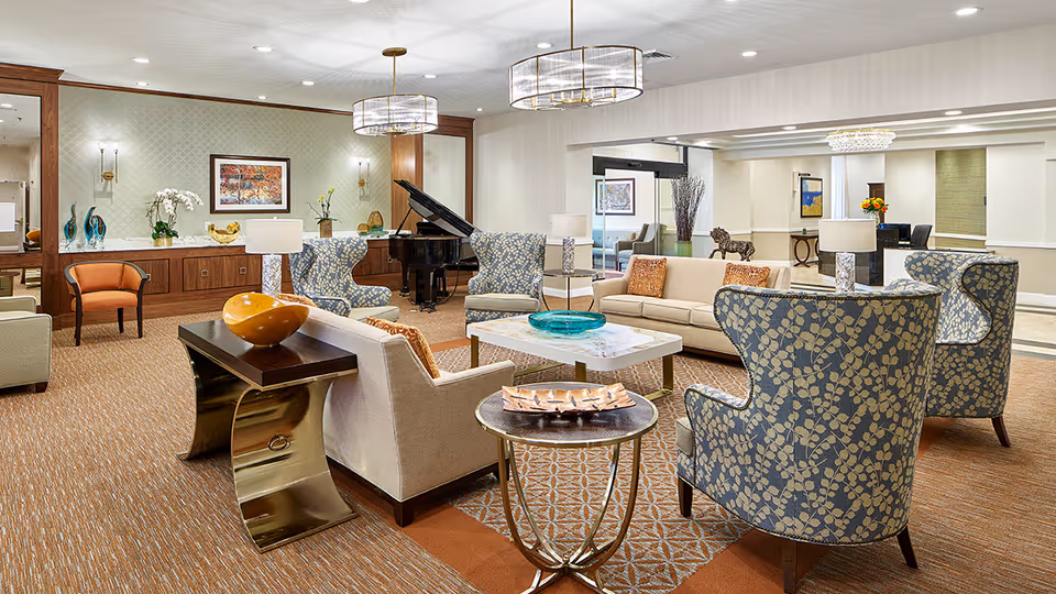A spacious and well-lit living room area in a senior living facility featuring multiple upholstered armchairs with floral patterns, beige sofas with orange cushions, a marble coffee table with a decorative blue bowl, side tables with lamps, a grand piano in the corner, and wall art. The room has warm carpeting and modern ceiling lights.