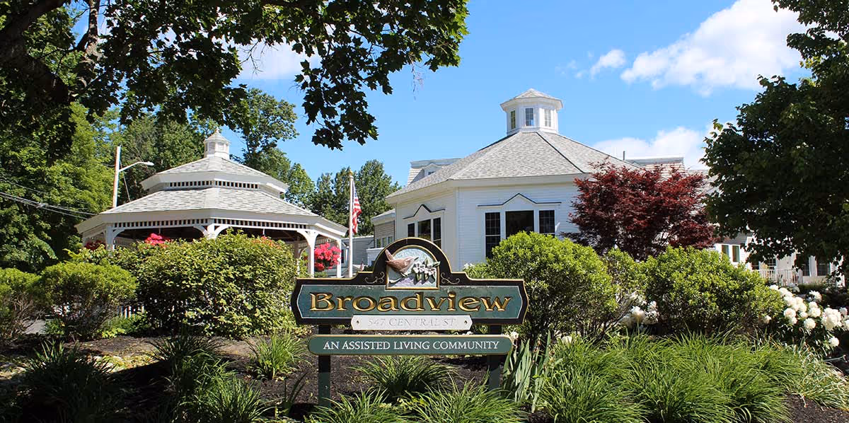 Exterior view of Broadview Assisted Living facility showing a white building with a cupola, a gazebo, lush green bushes, trees, and a sign in front that reads 'Broadview, An Assisted Living Community'.