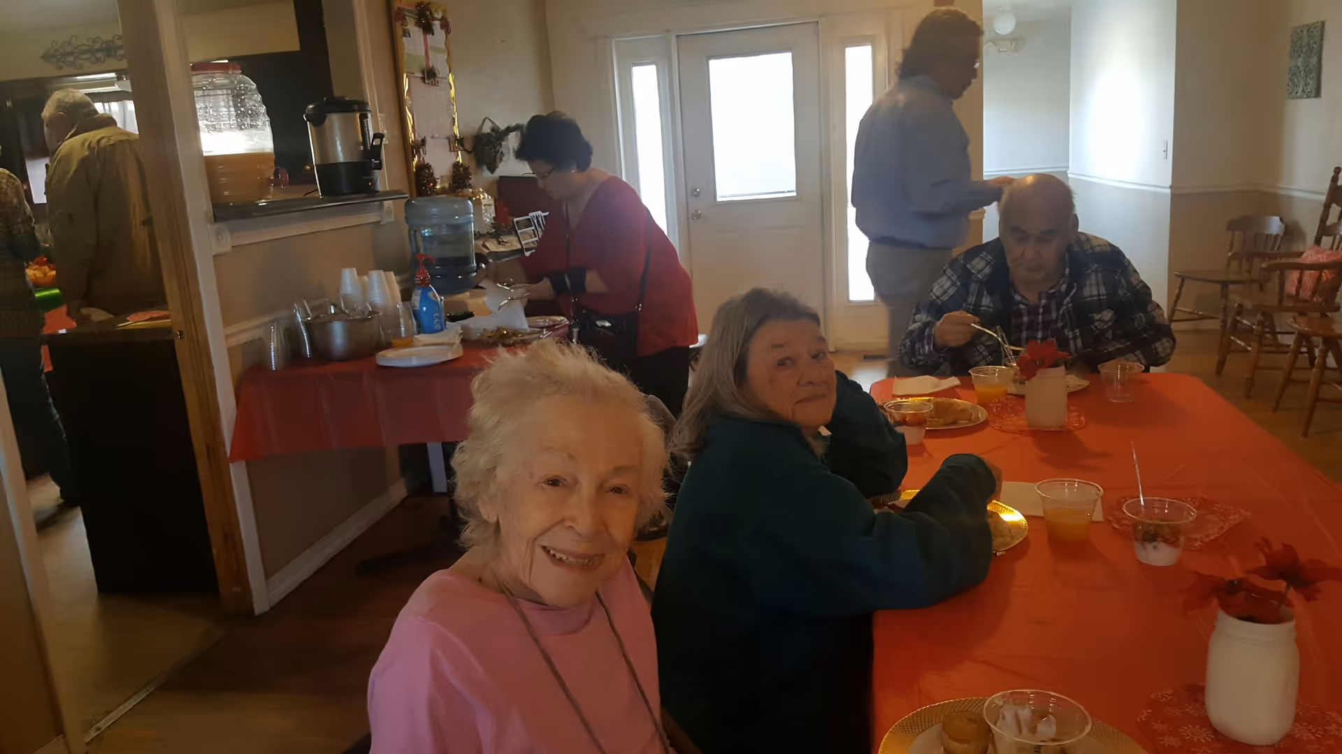 Several people sit and eat at a long table with an orange tablecloth in a communal dining area.
