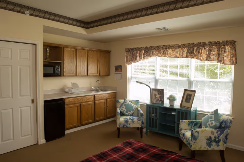 Sunlit studio-style living area with a small kitchenette, two patterned armchairs by a large window, and a bed partly visible in the foreground.