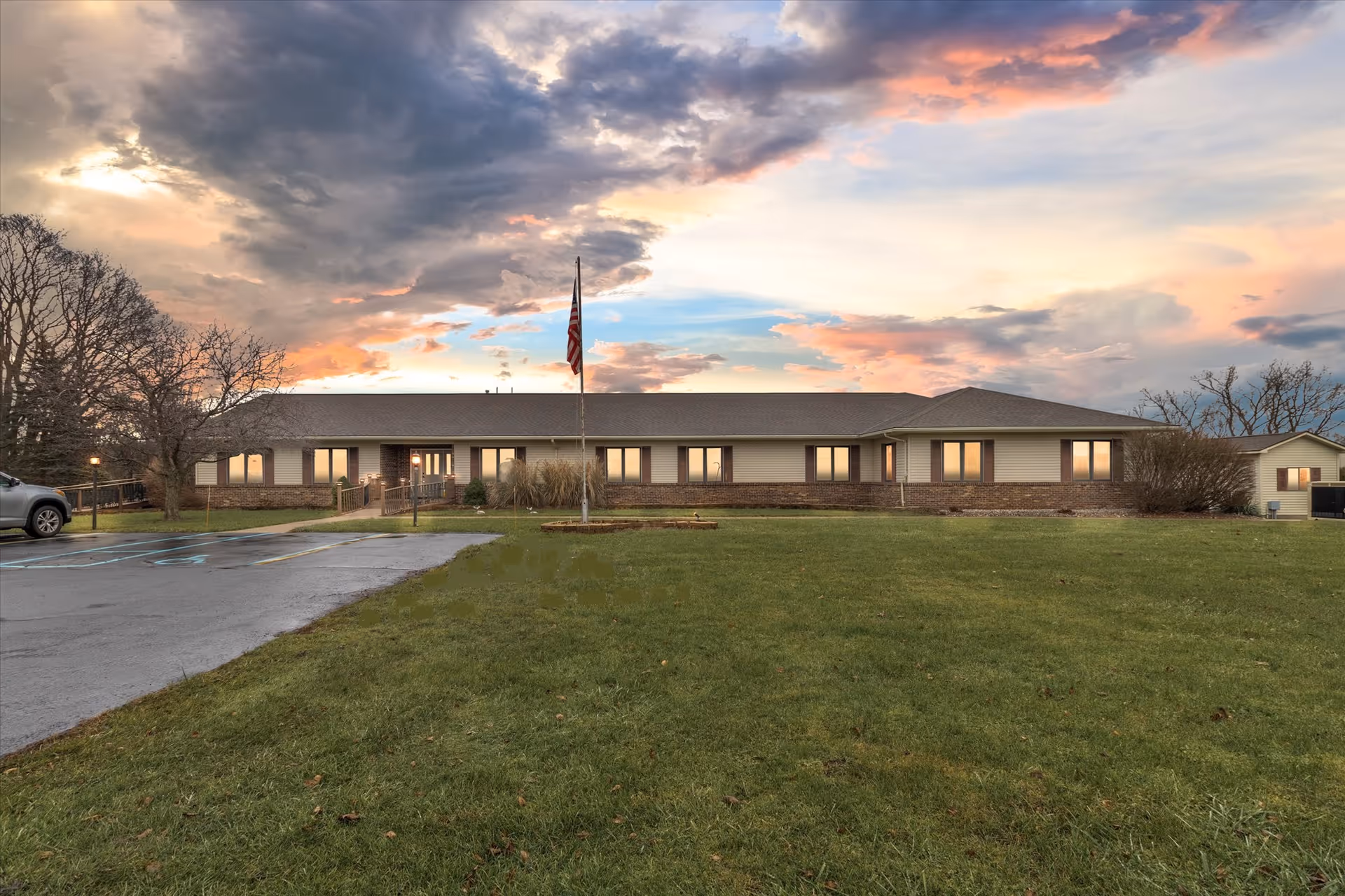 Exterior front view of a single-story building with multiple windows, a flagpole with an American flag in front, a parking area with a car on the left, and a large grassy lawn under a colorful sky at sunset.