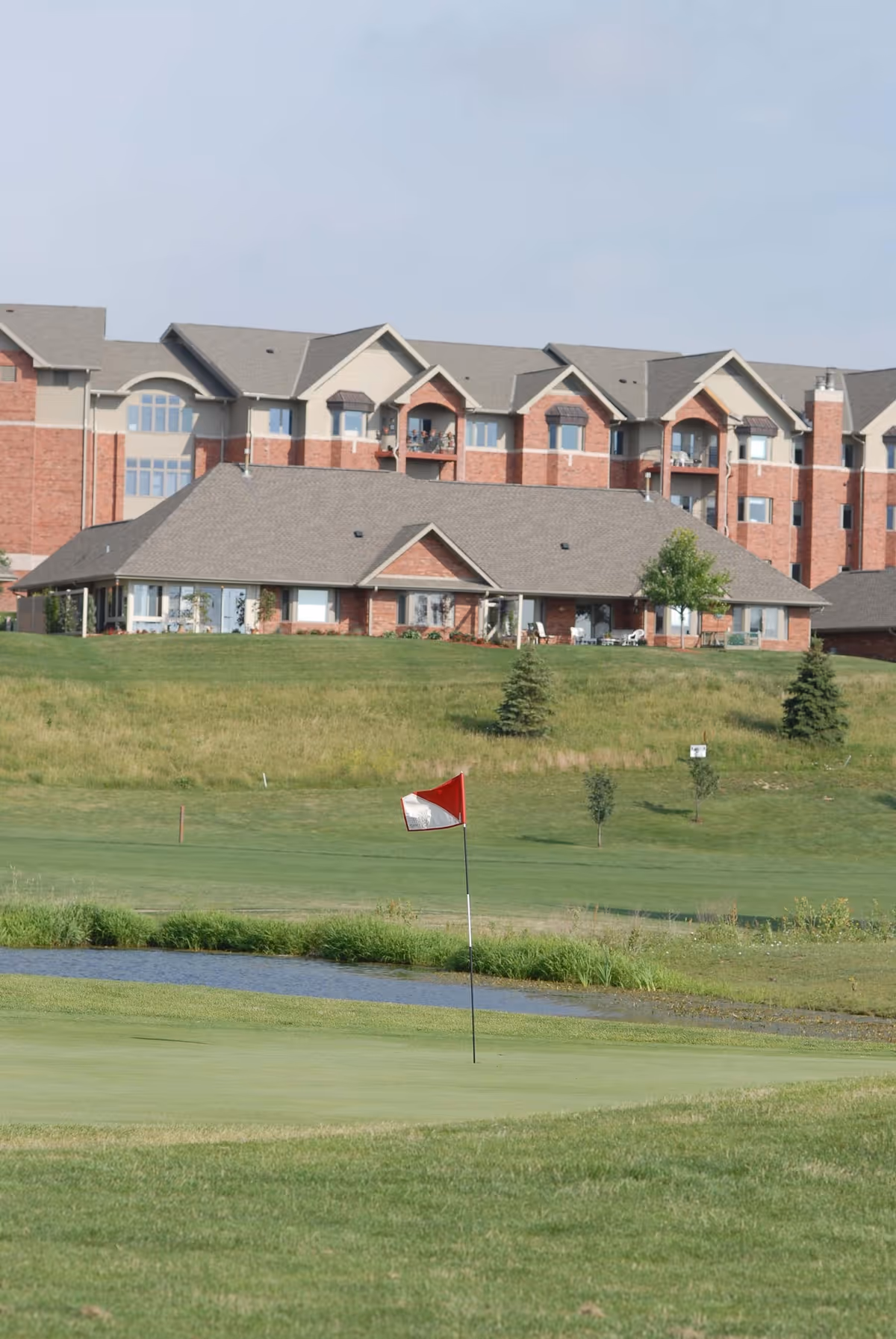 A large senior living facility building with multiple floors and balconies, situated behind a green golf course with a red and white golf flag on the putting green and a small water hazard.
