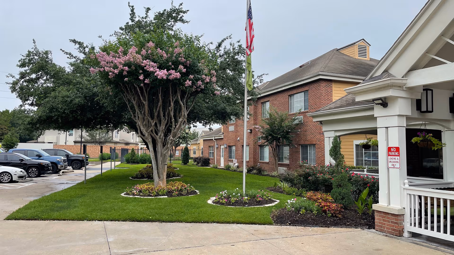 Exterior view of Atria Westchase senior living facility showing a well-maintained lawn with flower beds, a tree with pink blossoms, an American flag on a flagpole, a brick building with windows, and a covered entrance with a 'No Parking Loading & Unloading Only' sign. Several parked cars are visible on the left side.