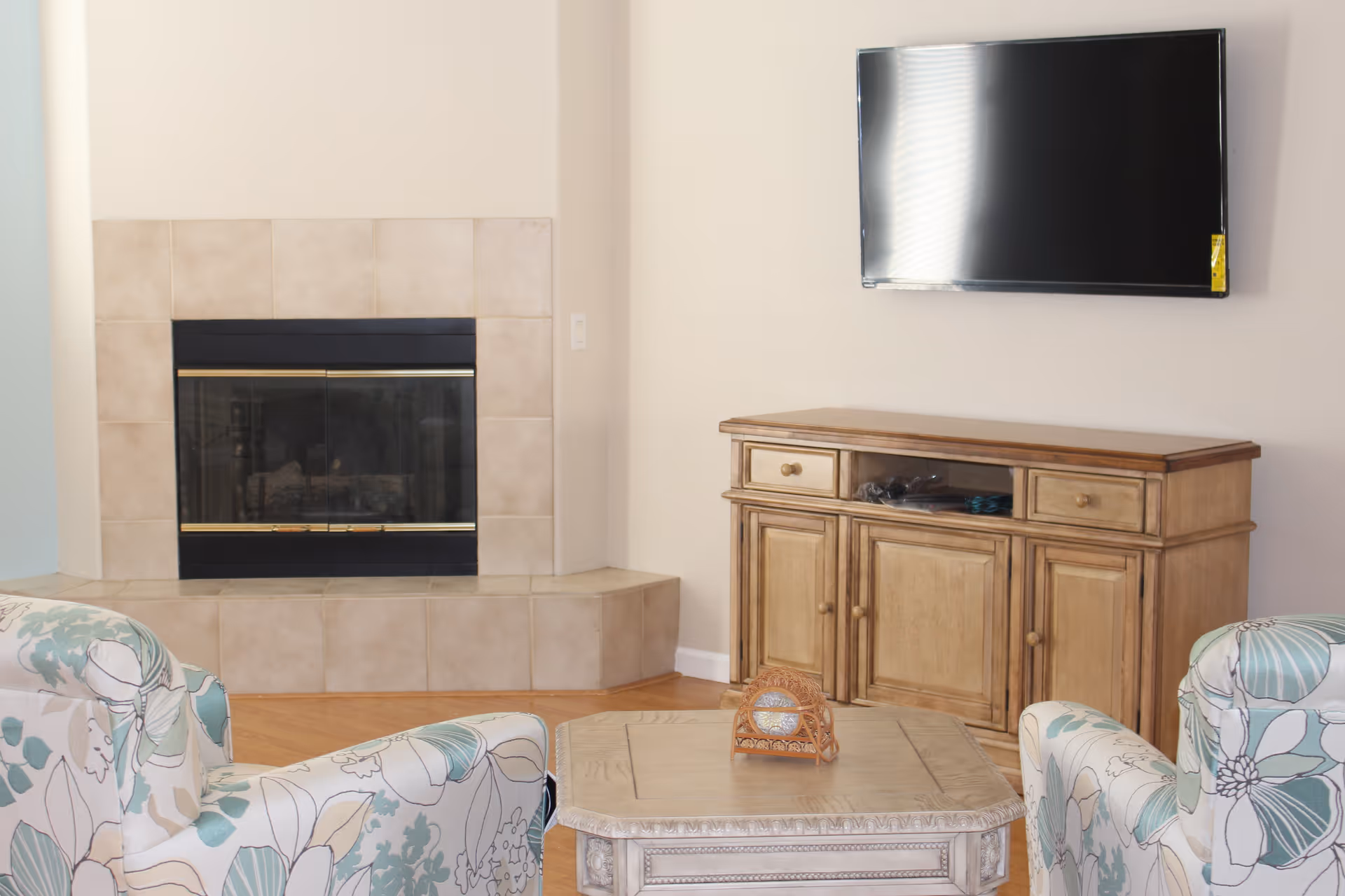 A cozy living room with a tiled corner fireplace, a wooden TV stand with a flat-screen TV mounted above it, and two floral patterned armchairs facing a wooden coffee table.