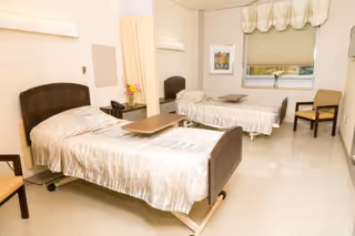 A clean and well-lit healthcare facility room with two hospital beds, each with light-colored bedding and a small overbed table. There is a nightstand with a telephone and a vase of flowers between the beds. A window with a valance and a chair are visible on the right side of the room.