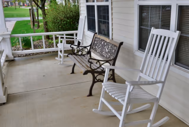 Covered front porch with white rocking chairs and a decorative metal bench overlooking a lawn.