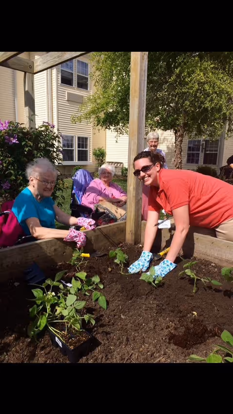 Three elderly women and a caregiver are gardening together in a raised garden bed outside a senior living facility. The caregiver, wearing a red shirt and blue gardening gloves, is planting seedlings in the soil while the elderly women, two seated in wheelchairs and one standing, watch and participate. The background shows the exterior of the facility with windows, trees, and flowering plants.