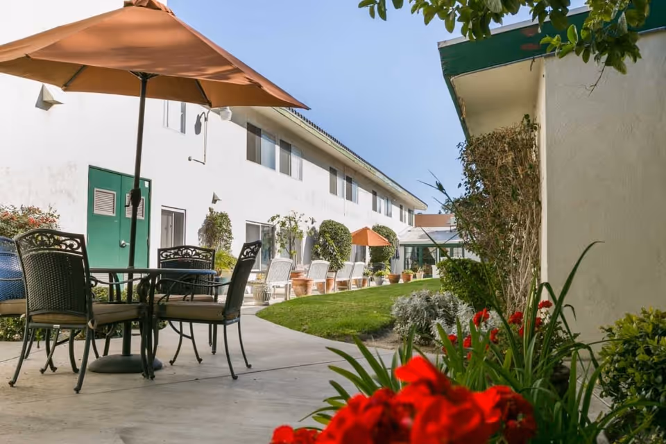 Outdoor patio area at Mainplace Senior Living with a round table and four chairs under a large orange umbrella. The patio is adjacent to a white building with green doors and windows. There are potted plants and neatly trimmed bushes along the building, with a well-maintained grassy area and red flowers in the foreground.