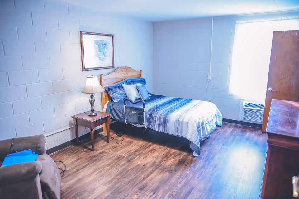 A simple bedroom with a single bed covered in blue and gray striped bedding, a wooden headboard, and several pillows. Next to the bed is a wooden nightstand with a lamp. There is a framed picture hanging on the white brick wall above the nightstand. A brown armchair with a blue item on it is partially visible on the left side. The room has wooden flooring, a window with bright light coming through, and a wooden door. An air conditioning unit is installed below the window.