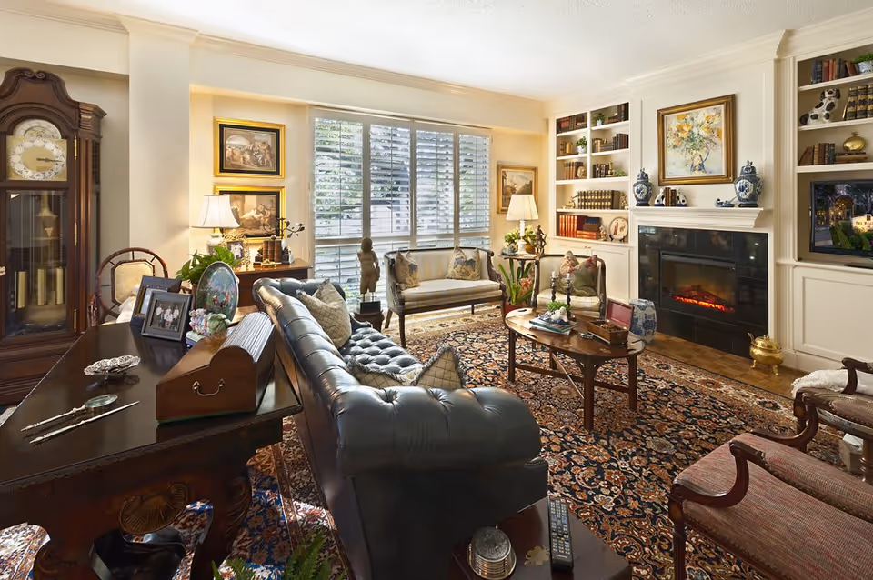 A cozy living room with a dark leather tufted sofa, antique wooden desk with framed photos, a grandfather clock, and a patterned area rug. The room features built-in bookshelves filled with books and decorative items, a fireplace with blue and white vases on the mantel, and large windows with white plantation shutters letting in natural light.