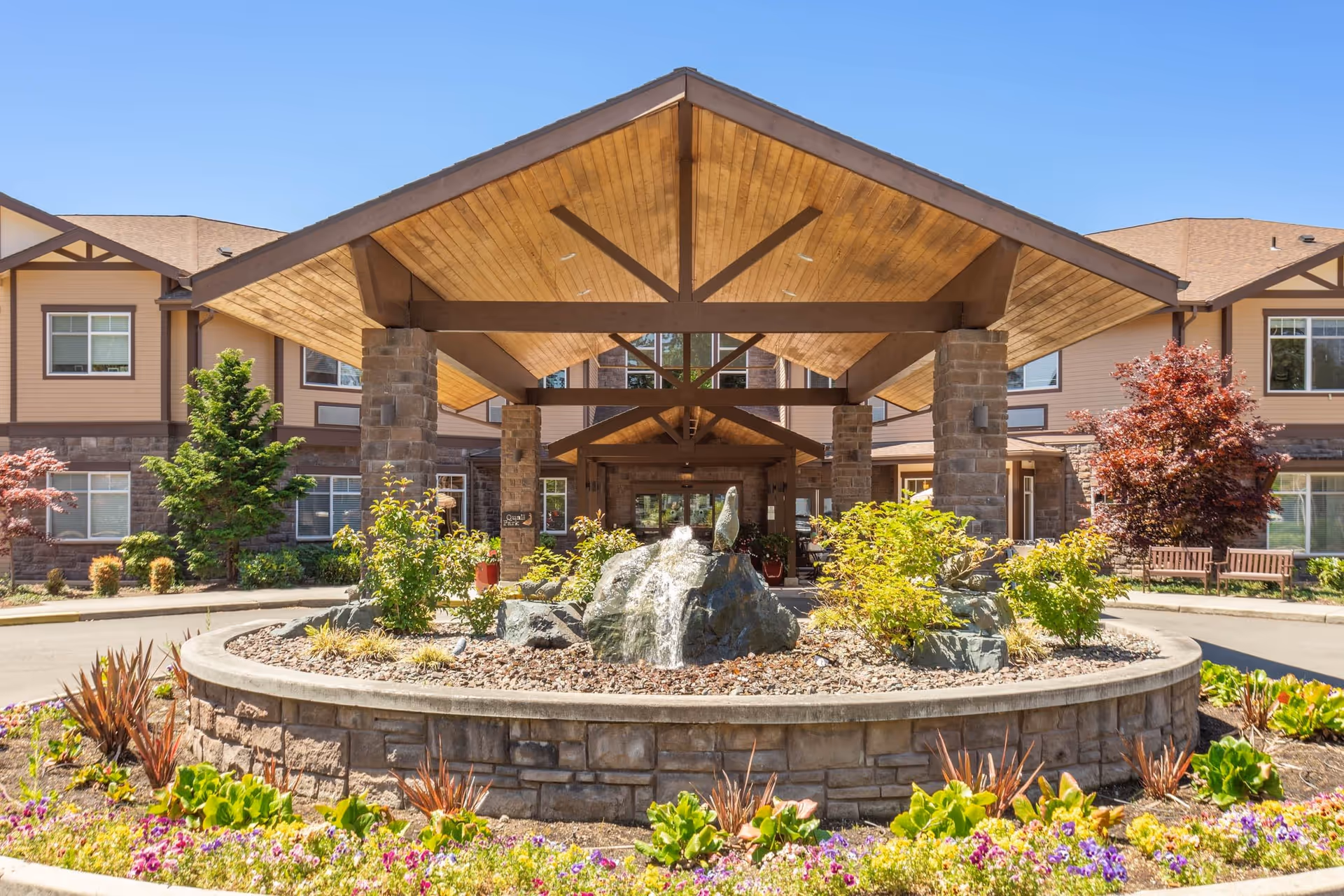Front entrance of Quail Park of Lynnwood senior living facility with a covered driveway supported by stone pillars, a circular stone planter with greenery and a rock fountain, and a landscaped garden with colorful flowers under a clear blue sky.