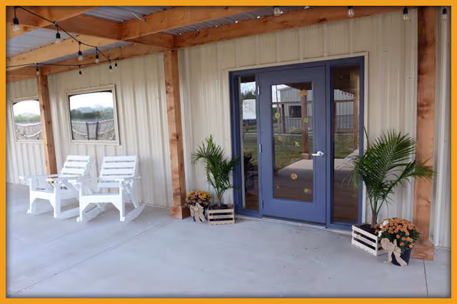 Covered outdoor patio area with two white wooden chairs on the left side, two potted plants with flowers placed symmetrically on either side of a blue double door entrance, beige siding walls, and string lights hanging from the wooden beams above.