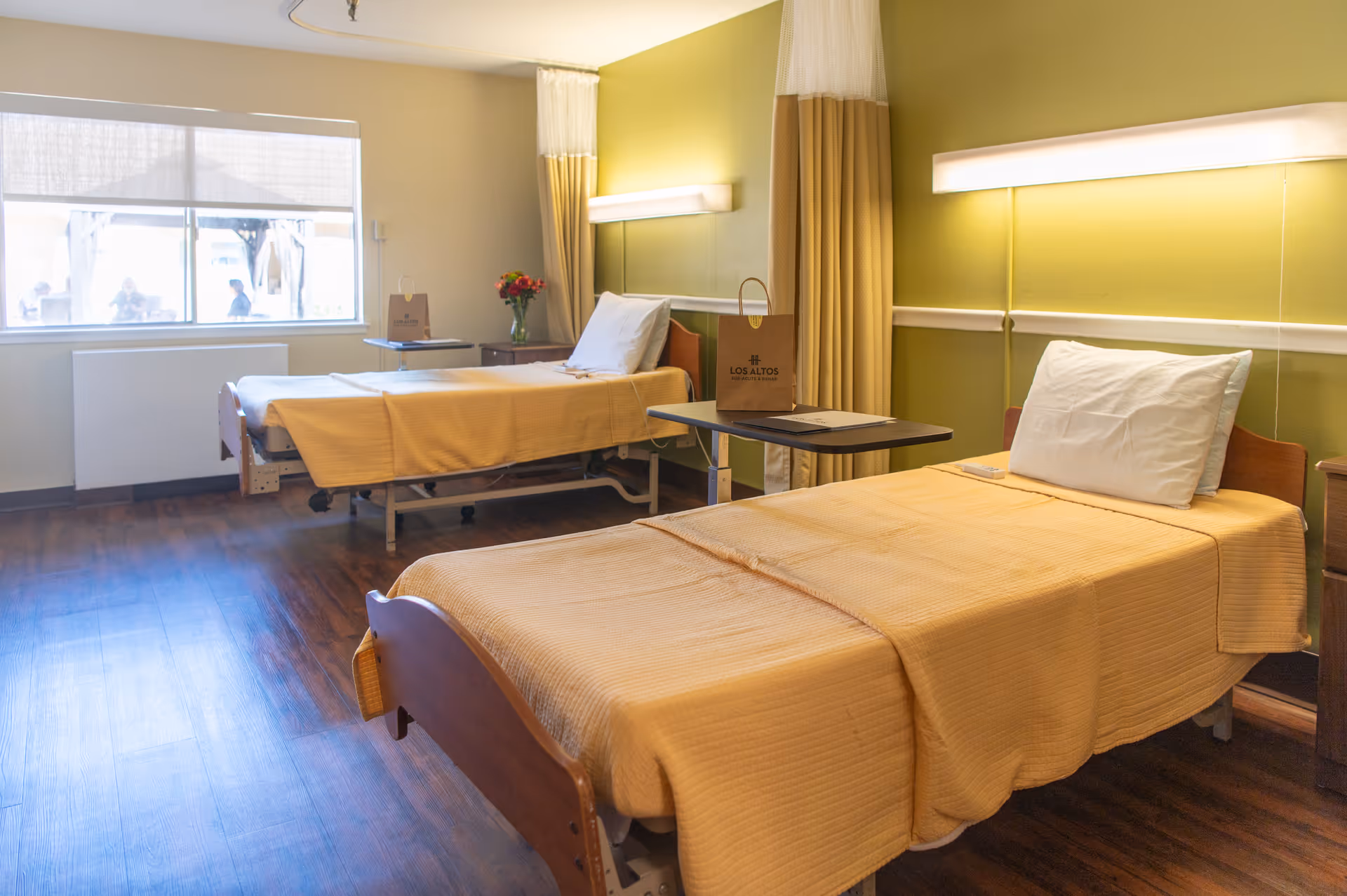 Two hospital-style beds with yellow bedspreads in a bright senior care room with a window and bedside tables.