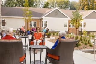 Several elderly people sitting and conversing on a patio area outside a residential building with trees and shrubs in the background.