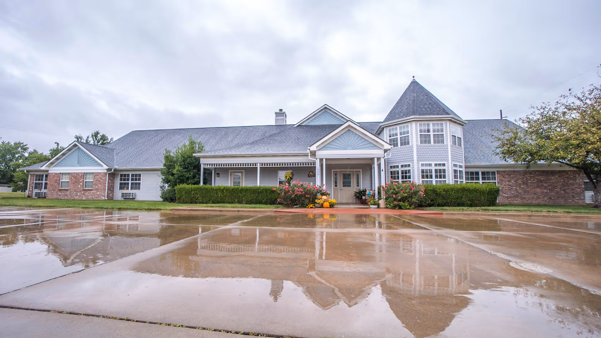 Front exterior of a single-story senior care building with a covered entrance, turret-style bay windows, landscaping, and a wet driveway reflecting the facade.