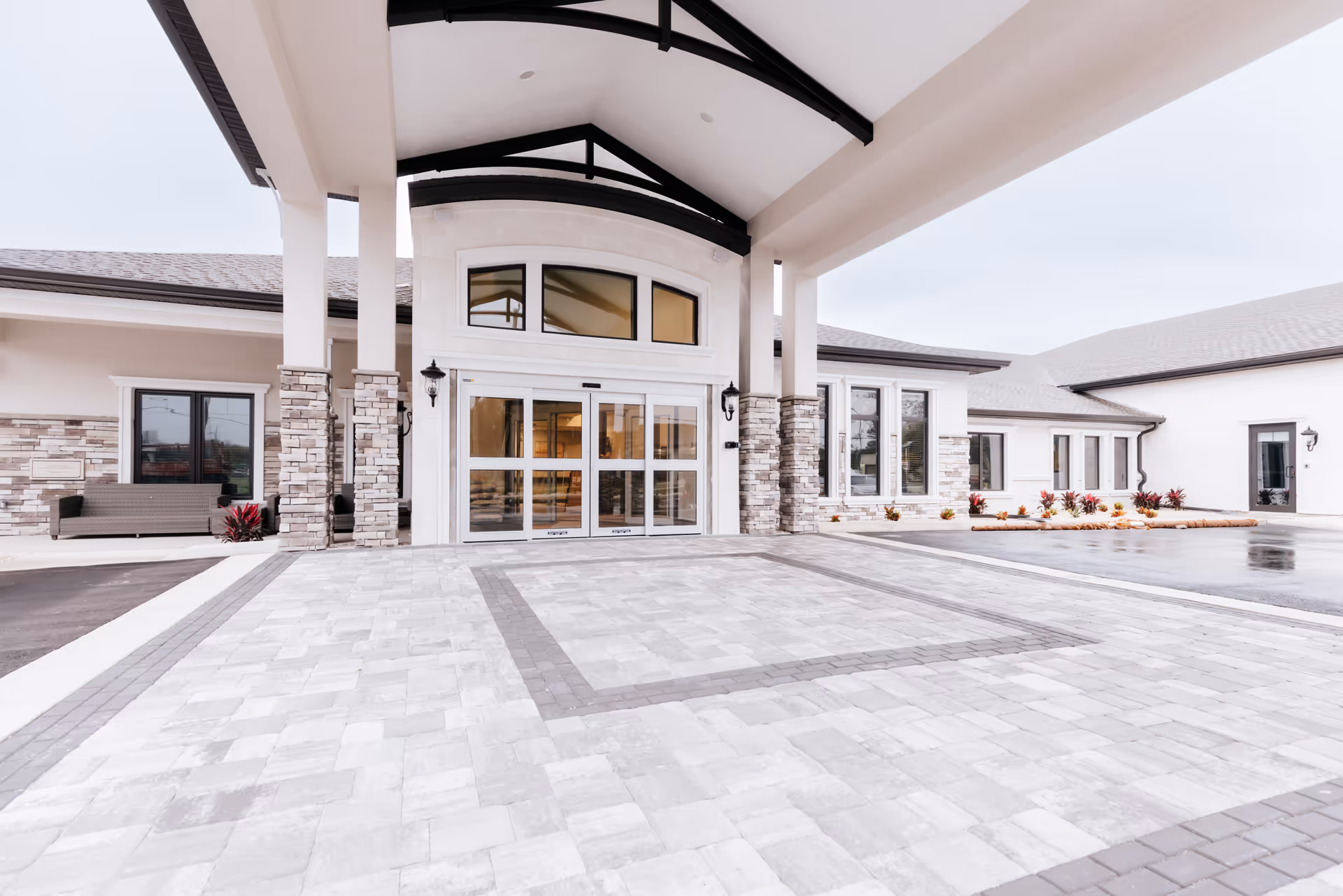 Entrance of a senior living facility with a covered drop-off area, automatic sliding glass doors, stone and brick exterior walls, and a paved driveway. There are benches and landscaped plants near the building.