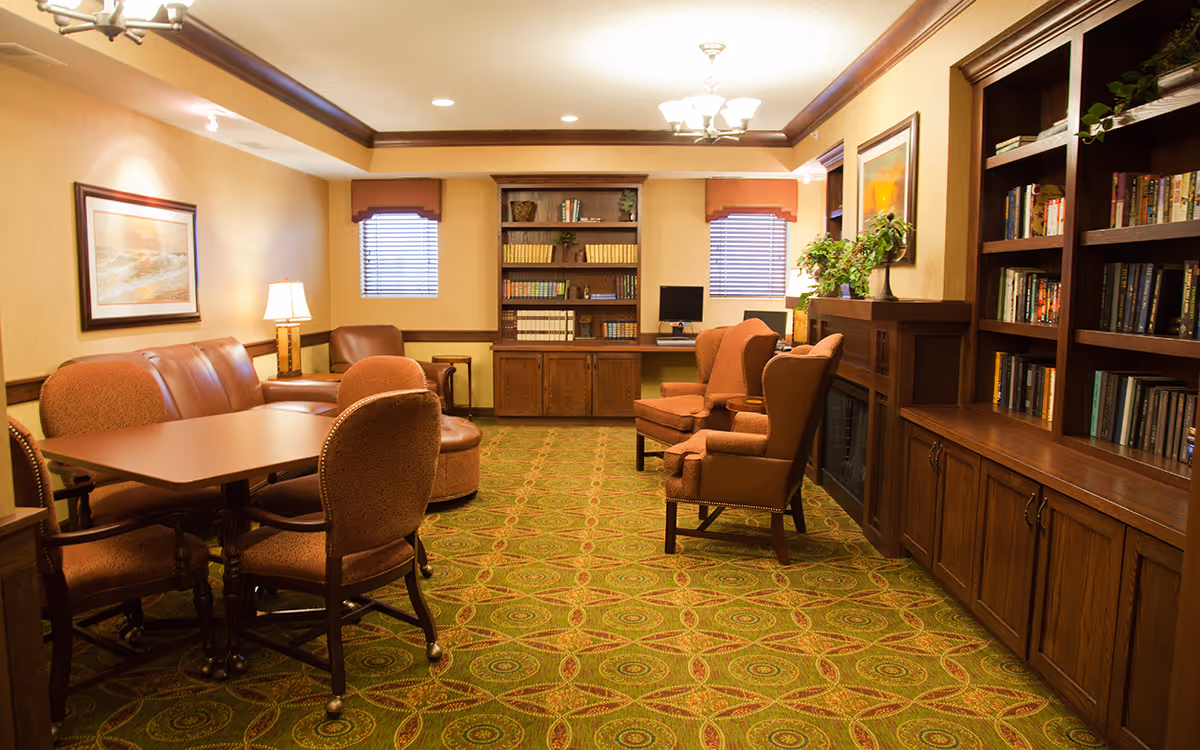 Cozy senior living common room with upholstered chairs, tables, bookshelves, and warm wood cabinetry.