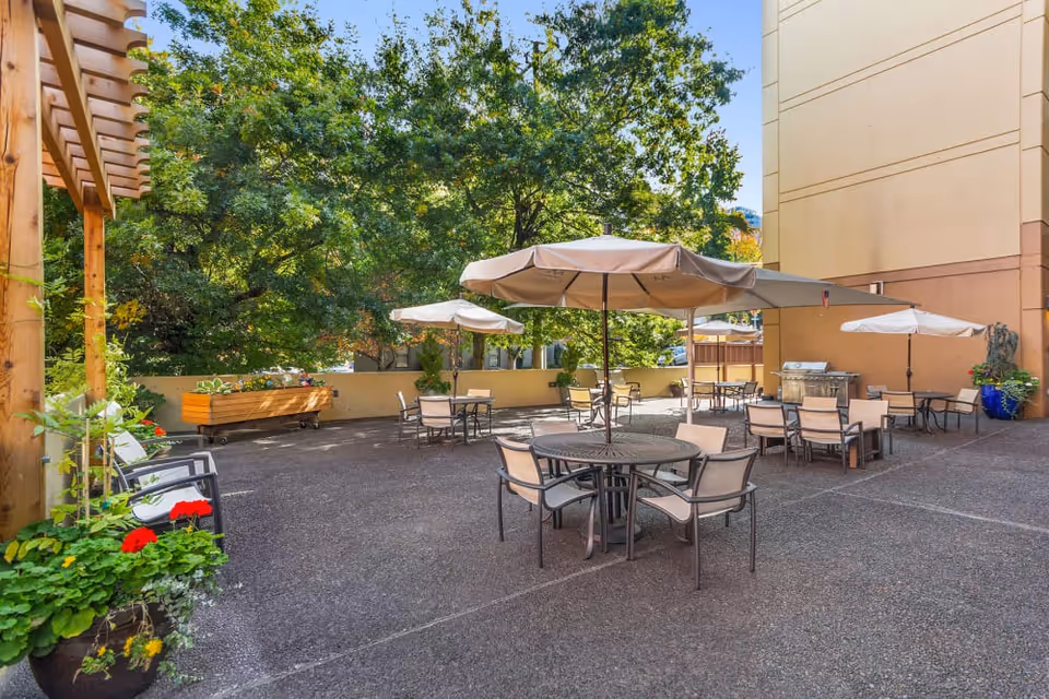 Outdoor patio area with multiple round tables and chairs, each shaded by large beige umbrellas. There are potted plants and flowers along the edges, a wooden pergola on the left, and a barbecue grill near the building wall. Trees and greenery are visible in the background.