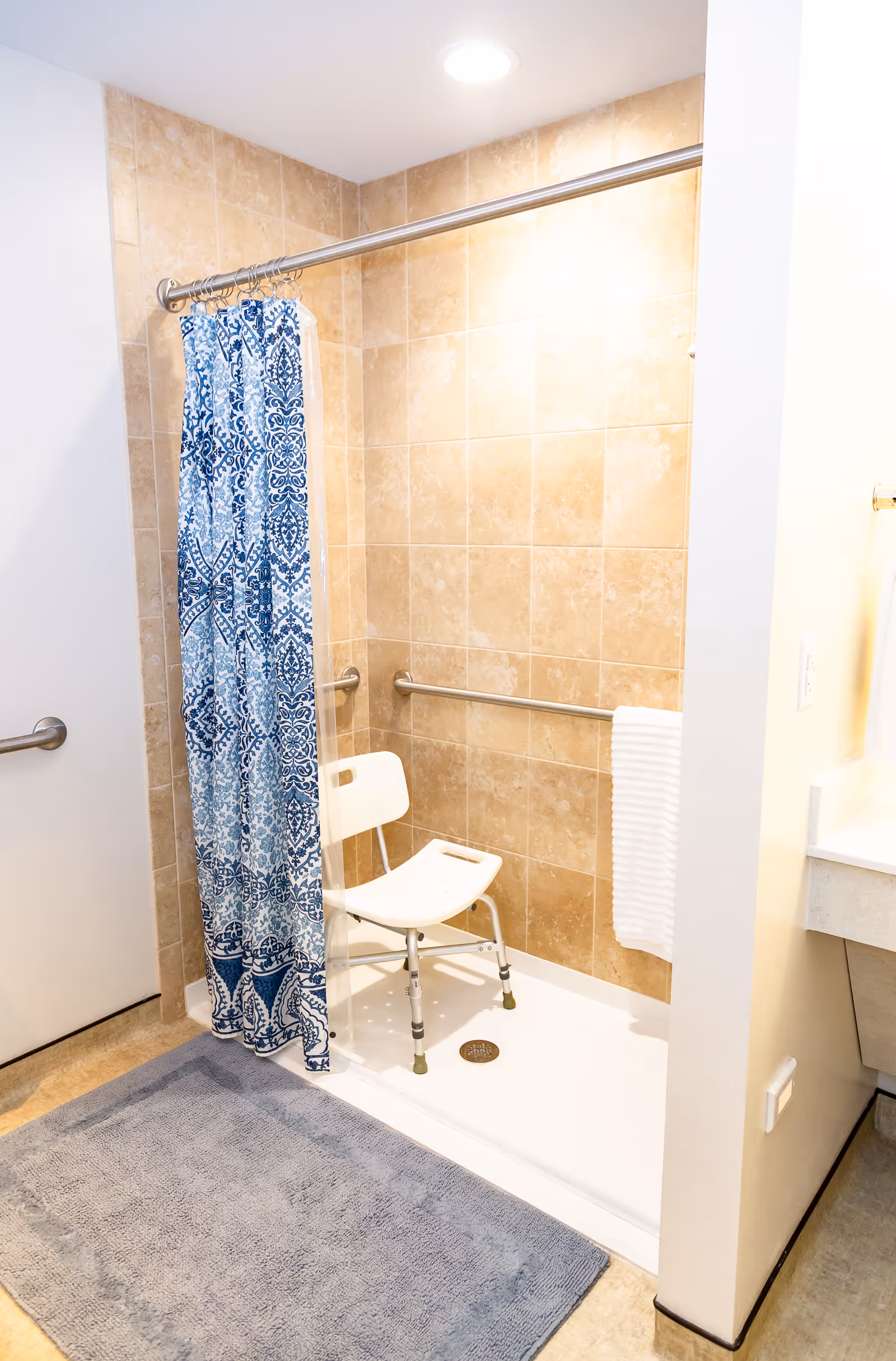 A bathroom shower area with beige tiled walls, a white shower chair, a blue and white patterned shower curtain, a gray bath mat on the floor, and a white towel hanging on a grab bar.