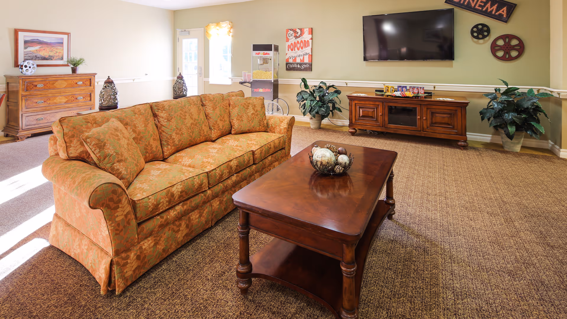 A cozy living room with a patterned orange sofa, a wooden coffee table with decorative spheres, a wooden TV stand with a flat-screen TV mounted above it, and two potted plants on either side. The wall behind the TV has cinema-themed decorations including a sign that says 'CINEMA' and film reels. There is a popcorn machine and a wooden dresser with a framed picture and a plant near a door and window letting in natural light.