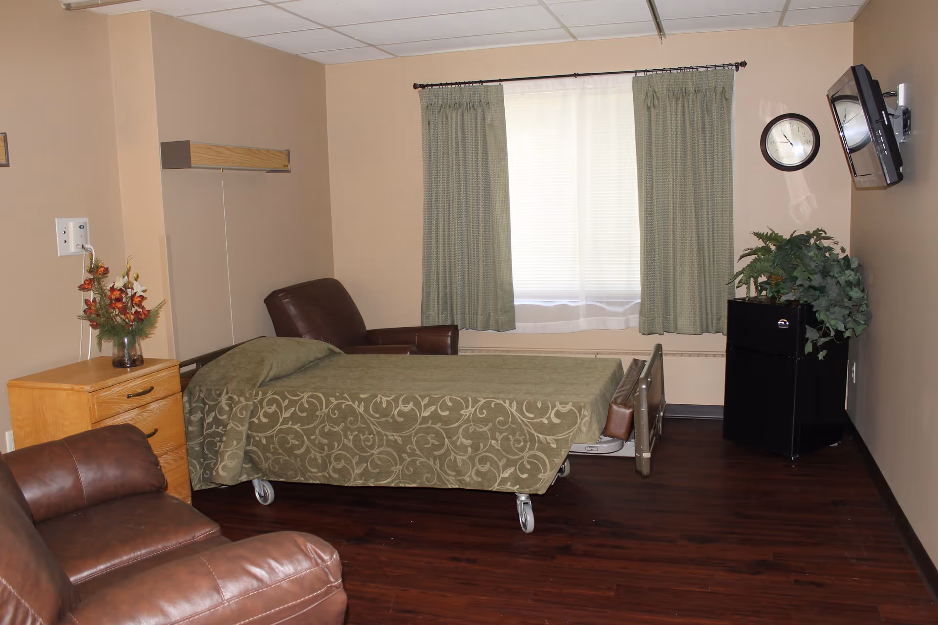 Private senior living bedroom with a single hospital-style bed, two leather chairs, a nightstand, wall-mounted TV, and a window with green curtains.