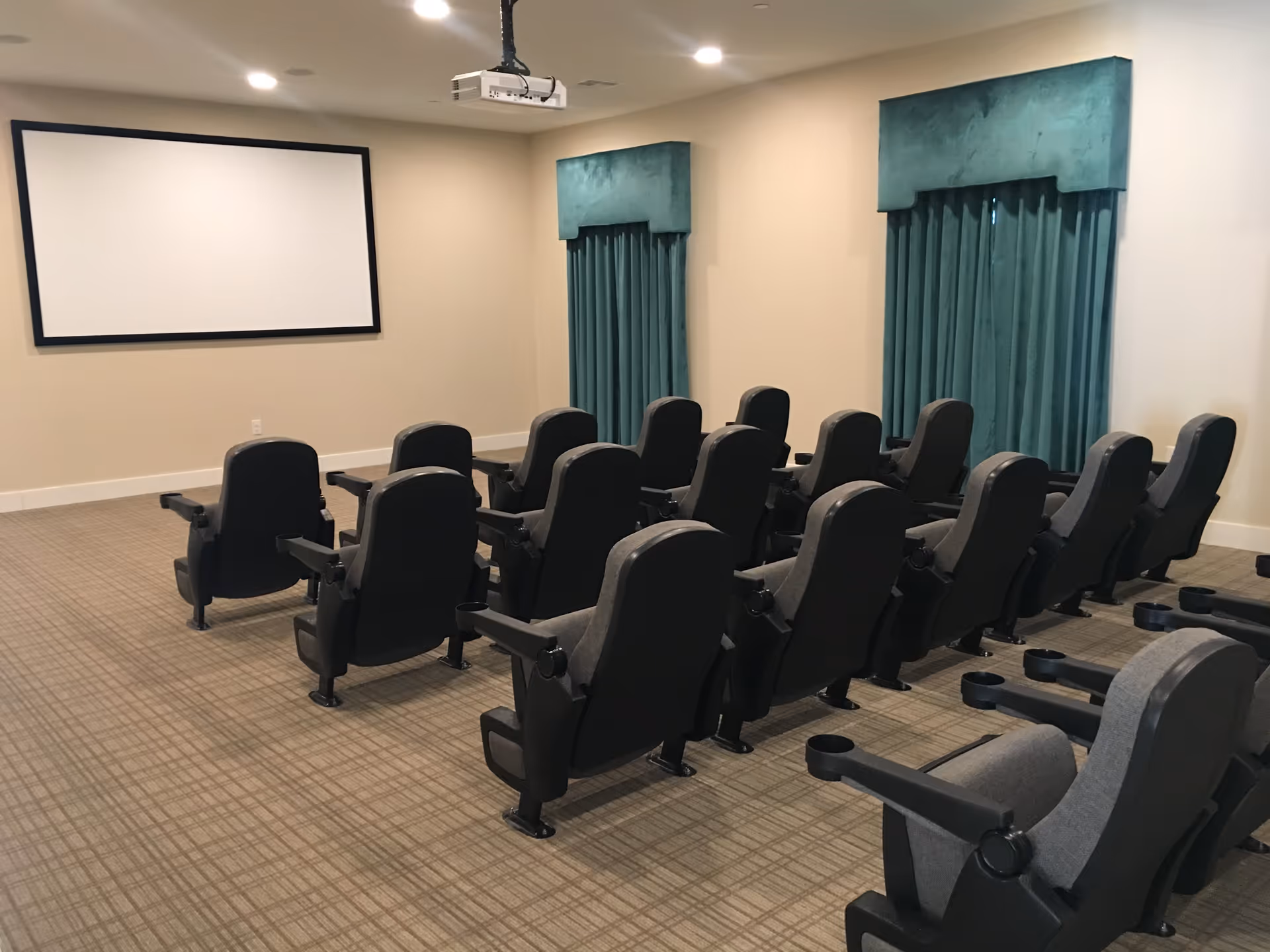 A small theater room with three rows of gray cushioned seats facing a large blank projection screen on a beige wall. The room has beige walls, carpeted floor, two windows with teal curtains, and a ceiling-mounted projector.