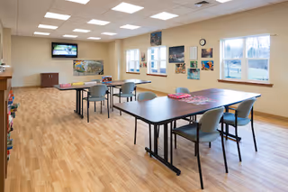 A bright and spacious room with wooden flooring, several tables and chairs arranged for group activities or dining. The walls have multiple colorful paintings and windows letting in natural light. A television is mounted on one wall above a cabinet.