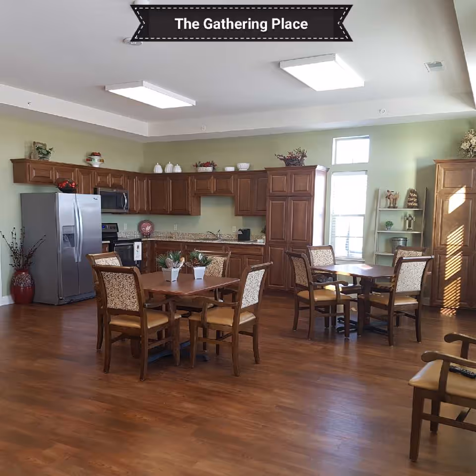 A spacious room with wooden flooring featuring a kitchen area with wooden cabinets, a stainless steel refrigerator, and a stove. There are two wooden dining tables each surrounded by four upholstered chairs. The room is well-lit with large ceiling lights and natural light coming through a window. Decorative plants and items are placed on top of the cabinets and shelves. A banner at the top reads 'The Gathering Place'.