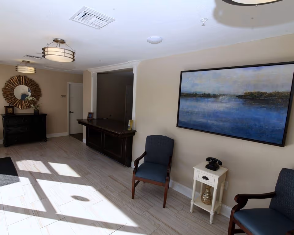 A well-lit interior space with two blue cushioned chairs, a small white side table with a vintage black rotary phone, a large framed painting of a serene water scene on the wall, a dark wooden counter, and a decorative round mirror above a black dresser. The floor is light-colored tile, and there are two ceiling light fixtures.