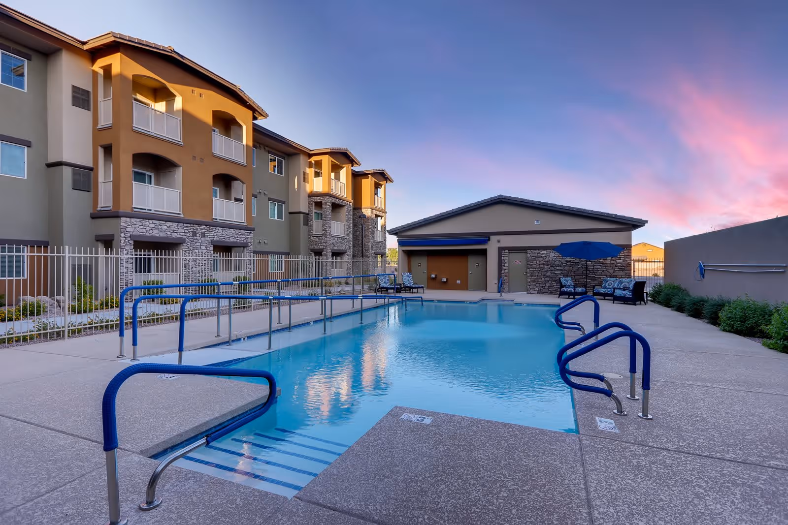 Outdoor swimming pool area at a senior living facility with blue handrails, lounge chairs, and a building with balconies in the background under a colorful sunset sky.