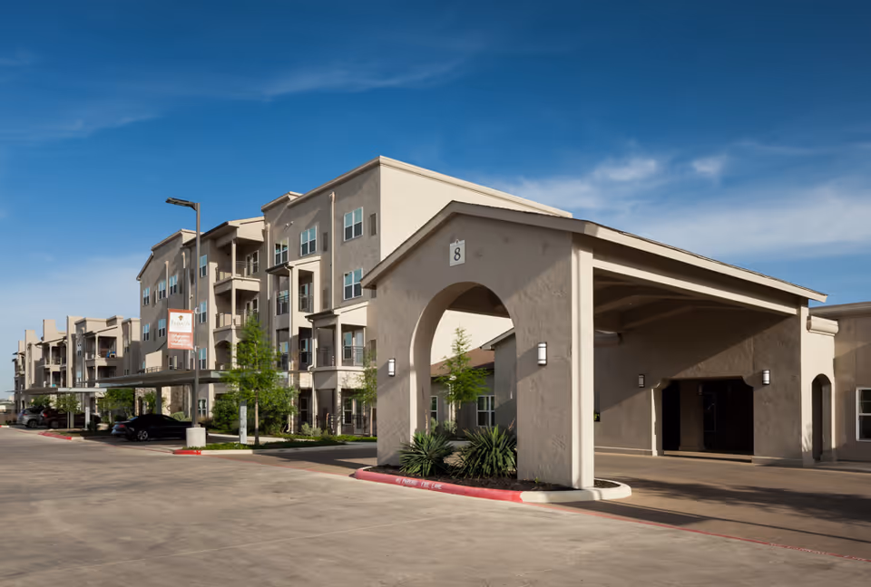 Exterior view of a multi-story senior living facility building with beige walls, balconies, and a covered entrance area under a clear blue sky. There are some parked cars and small trees along the driveway.
