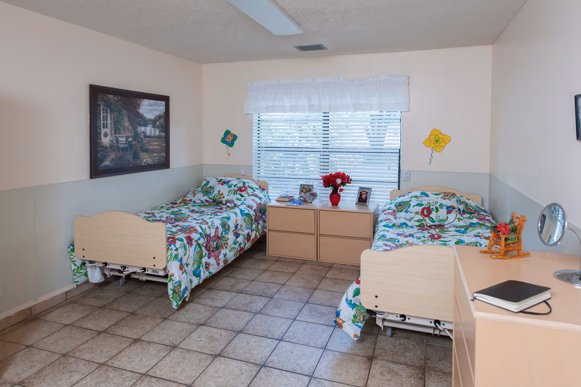 A bright room with two single beds featuring colorful floral bedspreads. Between the beds is a wooden dresser with framed photos and a vase of red flowers. A window with white blinds and a white valance is centered on the back wall. The room has tiled flooring and light-colored walls with simple flower decorations. A framed painting hangs on the left wall, and a small wooden dresser with a mirror and a notebook is on the right side.