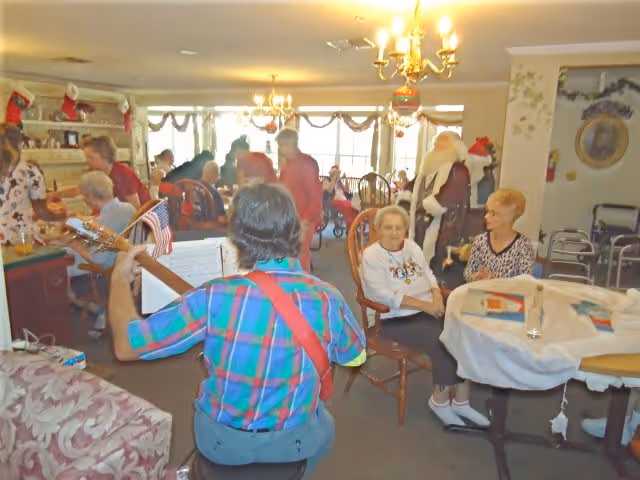 A group of elderly people and caregivers gathered in a decorated common room with Christmas stockings and garlands. A person in a plaid shirt is playing a guitar facing the group. Some residents are seated around tables and chairs, enjoying the festive atmosphere.