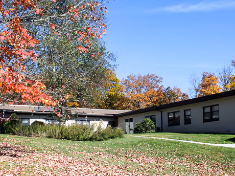 Single-story building with beige walls and dark trim surrounded by green grass and trees with autumn foliage under a clear blue sky.